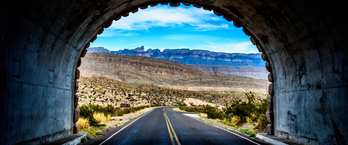 View from a tunnel framing a scenic desert road leading to distant mountains under a vibrant blue sky